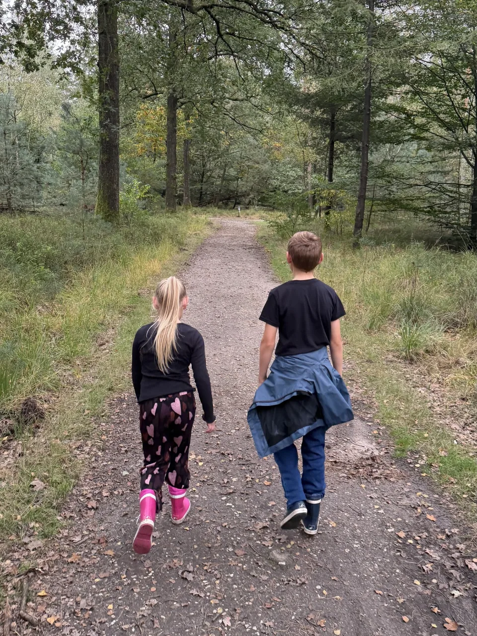 Twee kinderen in regenlaarzen lopen van achteren gezien naast elkaar over een bospad