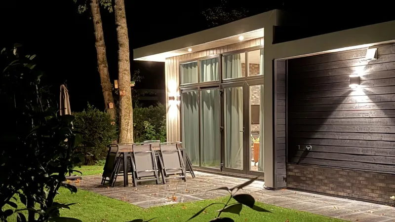 Het huis in de avond met verlichte glazen pui en buitenlampen, het terras met tuinset en het gazon