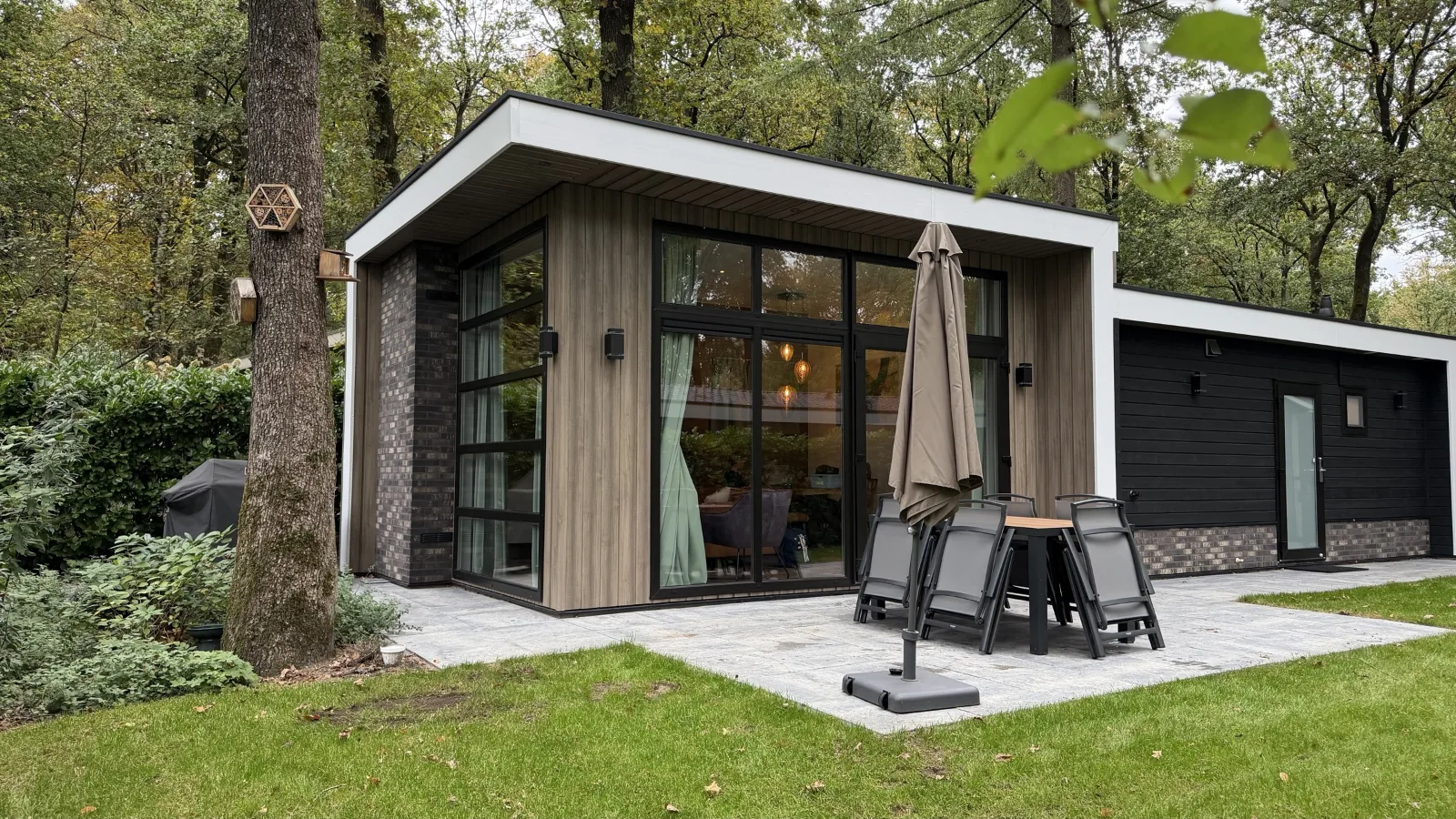 Het huis overdag vanaf de tuin: glazen pui, terras met tuinset en parasol, gazon en hoge bomen