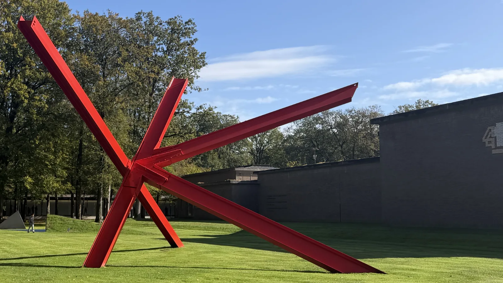 Groot rood stalen beeld van schuine balken op het gazon naast een museumgebouw, onder een blauwe lucht