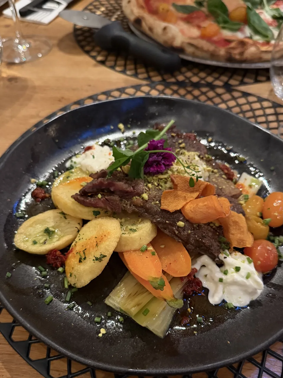 Hoofdgerecht met aardappeltjes, wortel en tomaatjes op een zwart bord bij De ProefTuin, met een pizza op de achtergrond