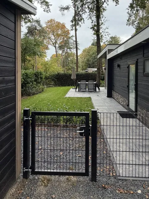 Het zwarte poortje met slot in het hek rond de tuin, met daarachter het gazon, het terras met tuinset en hoge bomen