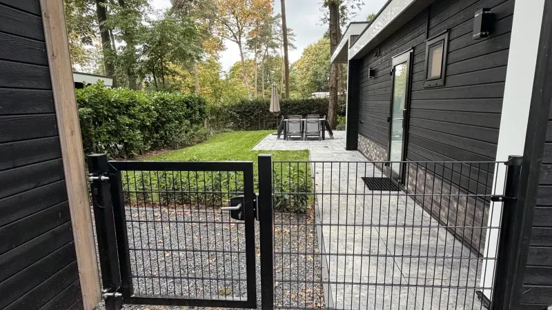 Zwart hek met poortje aan de zijkant van het huis, met daarachter het gazon, het terras met tuinset en parasol en een hoge haag